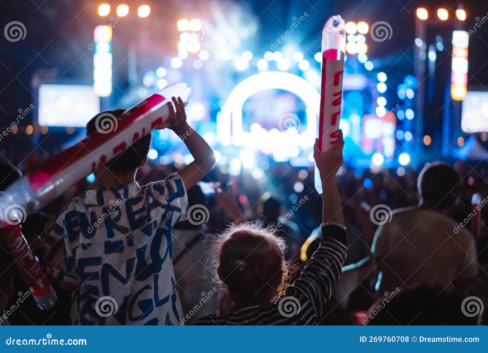 A performer on a concert stage with bright spotlights and an enthusiastic crowd raising their hands, holding microphones