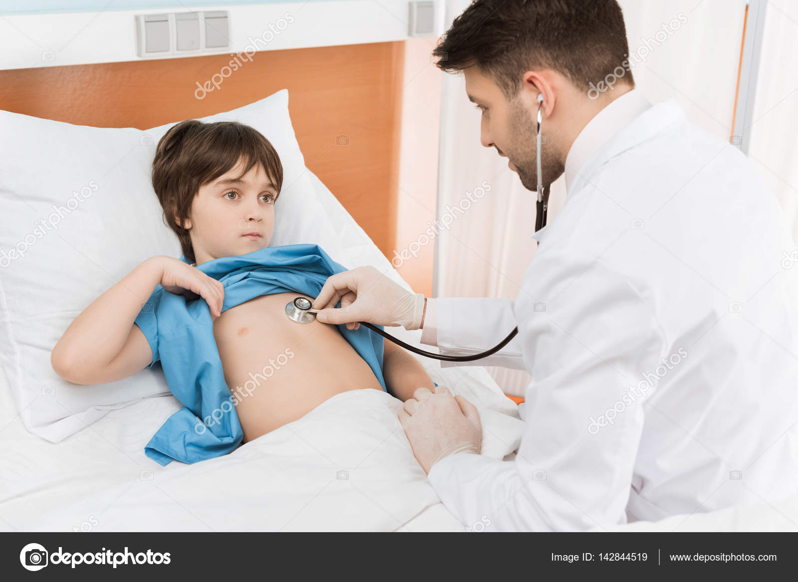 A doctor in a white coat gently examining a young child's throat in a brightly lit hospital examination room