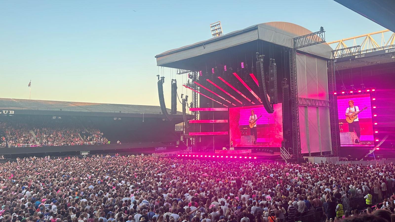 Ed Sheeran performing live on a massive concert stage with a crowd of thousands in the background, holding an acoustic guitar.