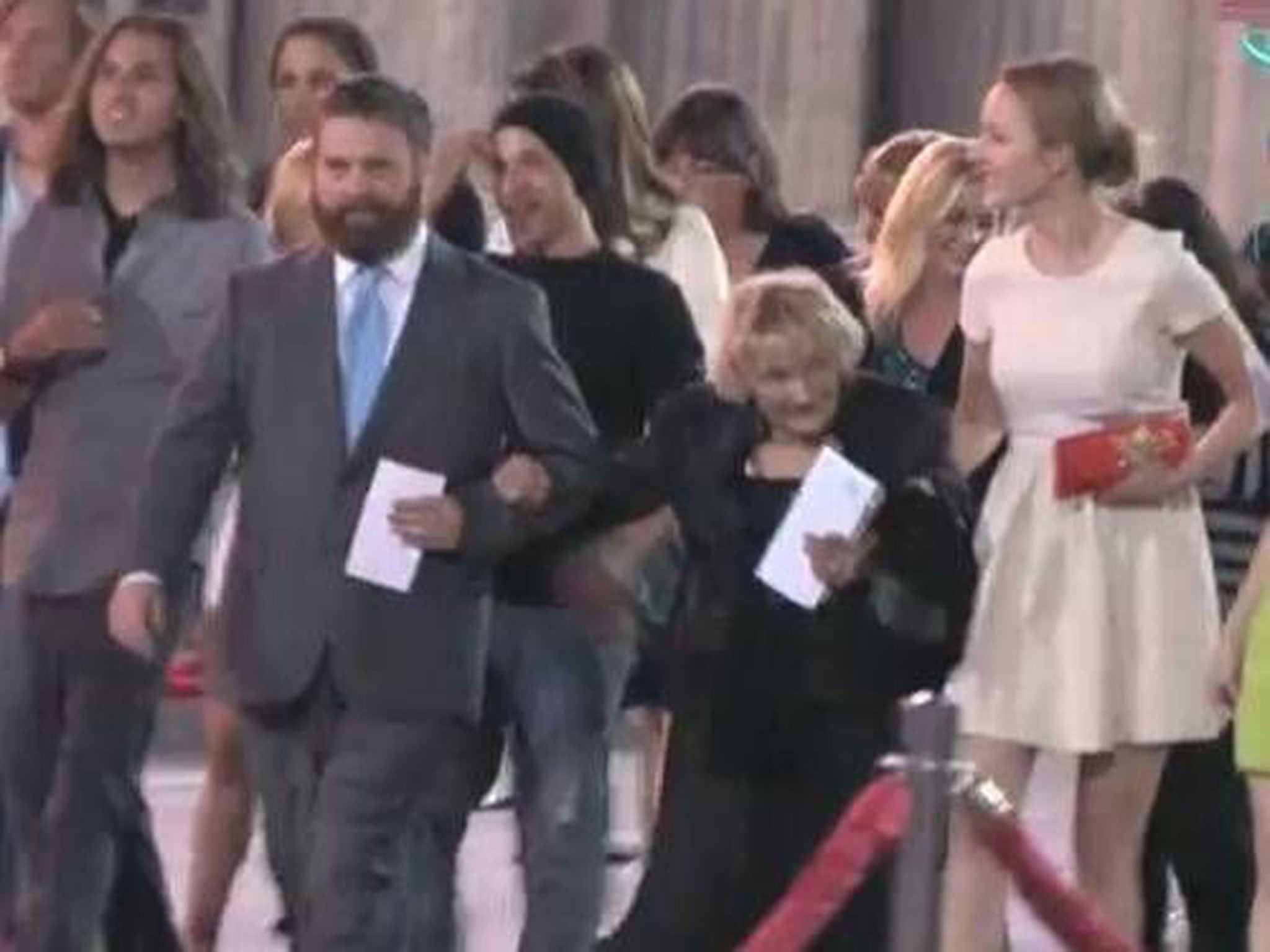 A male actor in a dark suit standing on a red carpet surrounded by photographers and fans