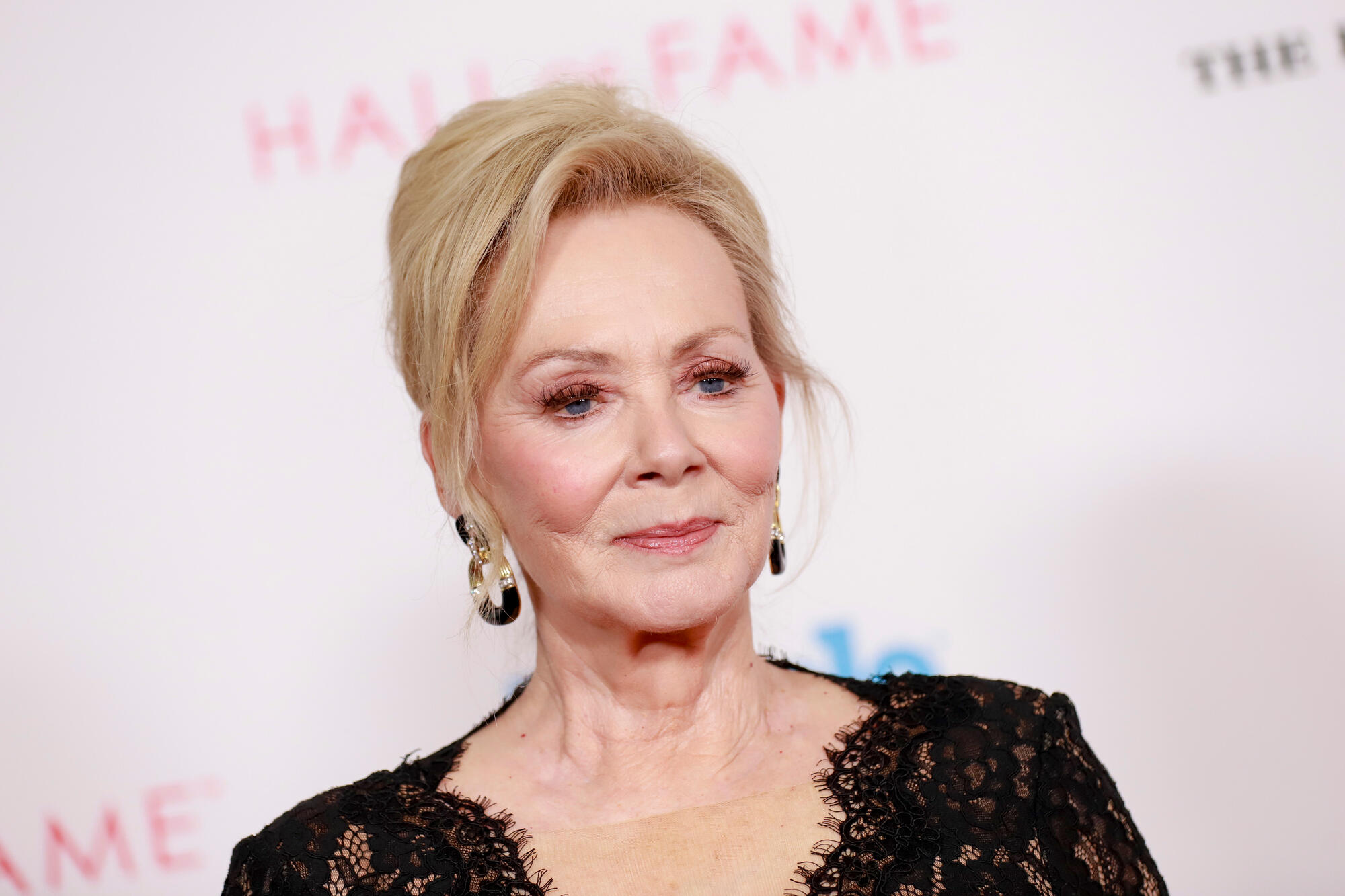 Jean Smart sitting on a stage in front of a microphone with warm lighting, wearing a stylish black outfit during an interview setting.
