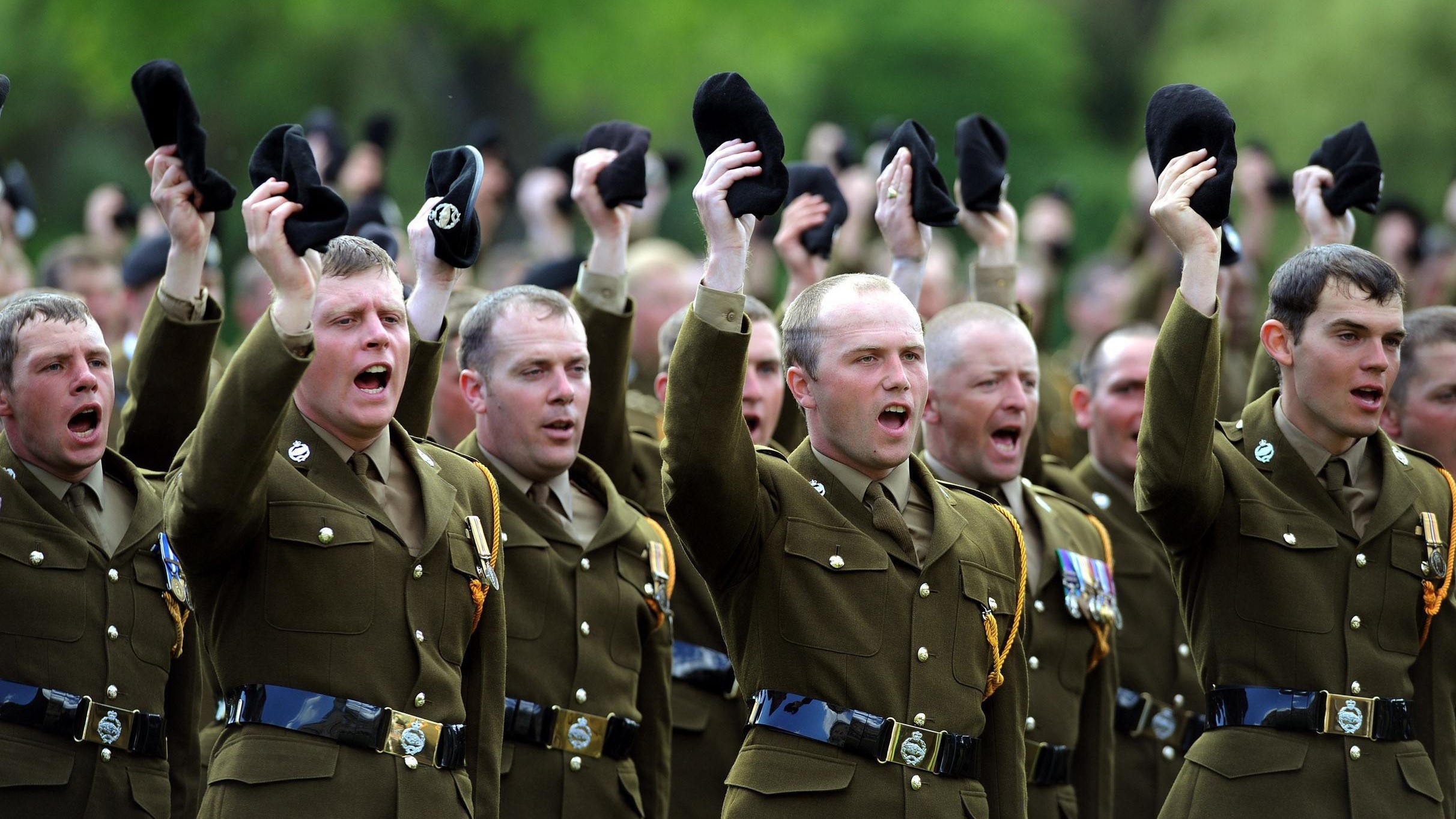 A line of British Army soldiers in ceremonial green uniforms marching in formation on a paved street with spectators in the background