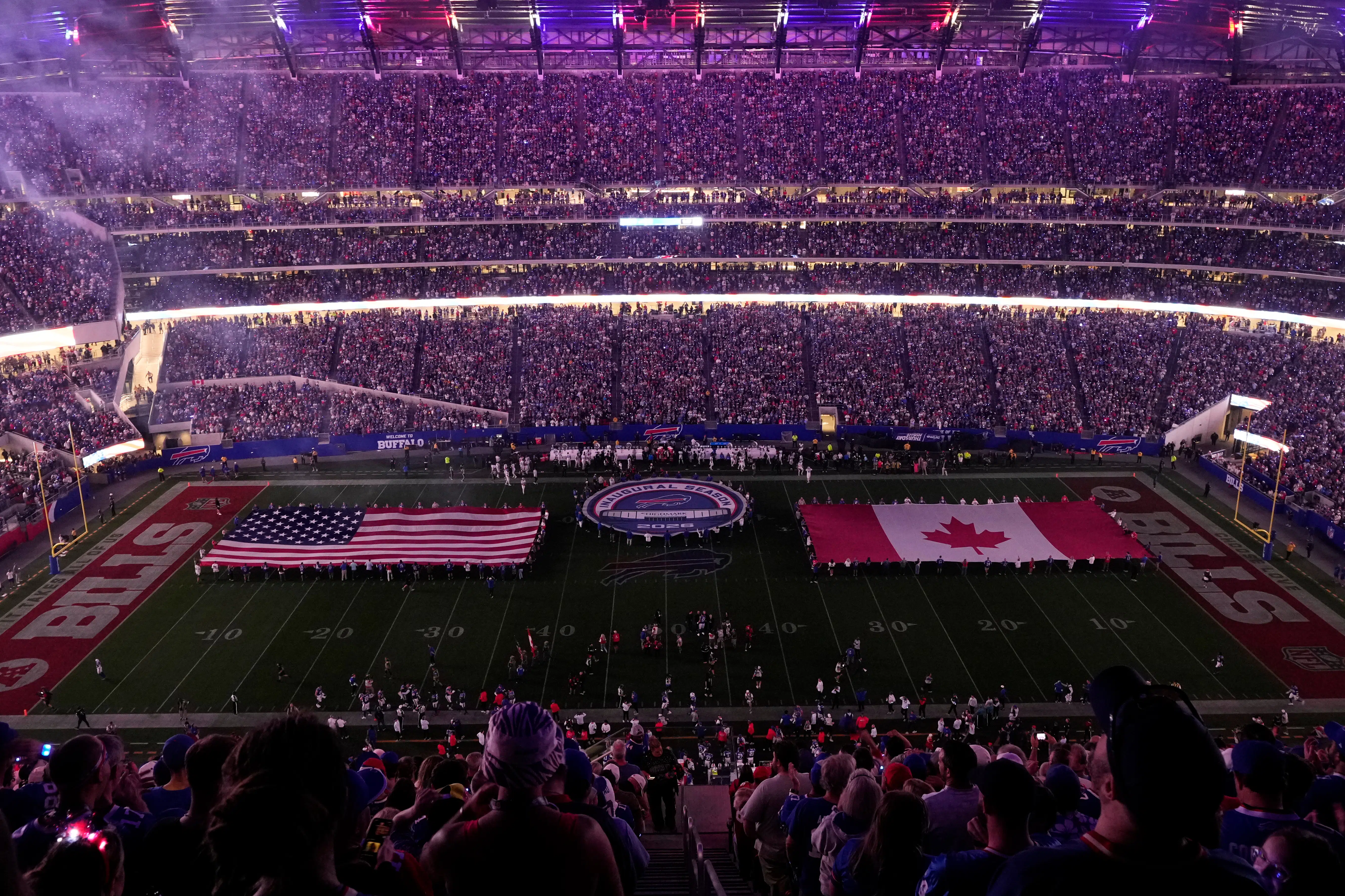 A packed American football stadium at night with bright floodlights illuminating the field and fans in team colors cheering in the stands