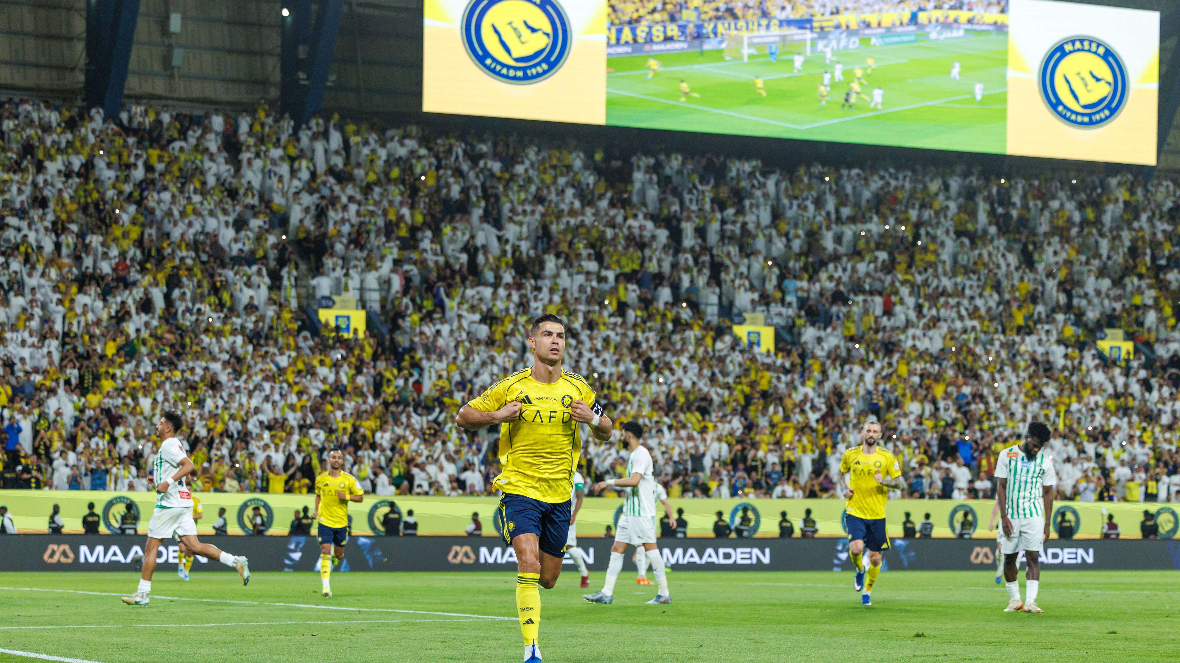 A packed soccer stadium with fans waving flags and cheering during an intense match between Al Ain and Al-Nassr